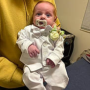 Scot joined the competition — help win amazing prizes! baby, infant, pacifier, white_suit, bow_tie, boutonniere, surprised, wide_eyes, person, hand, adult_arm, hoodie, beard, indoor, bedroom, crib, window_blinds, sitting, portrait, cute