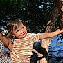 toddler, child, striped_clothing, tree, water, outdoor, nature, sunlight, shadow, person, holding, smile, curious, arm, river, leaves, baby, summer, daylight, playful