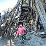 child, toddler, smiling, pink_hoodie, camo_pants, barefoot, sand, beach, driftwood, wood_structure, outdoor, nature, sunlight, playful, happy, portrait, tree_branches, daylight, person, fun