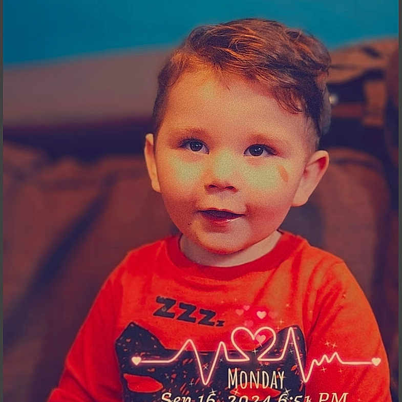 Alexandre is registered to the contest to win money with this photo: child, toddler, red_shirt, face, smile, couch, indoor, hair, portrait, young_child, sitting, cute, happy, person, digital_overlay, timestamp, heartbeat_graphic, blue_wall, cozy, casual