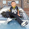 child, boy, portrait, outdoor, car, vehicle, football_jersey, steelers, sportswear, sneakers, black_clothing, yellow, brick_building, tree, driveway, sitting, smiling, short_hair, reflection, fashion