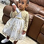 brown_sofa, carpet, child, curious, cute, dress, floor, girl, gold, indoor, living_room, person, portrait, remote_control, shoes, small, socks, standing, white, young