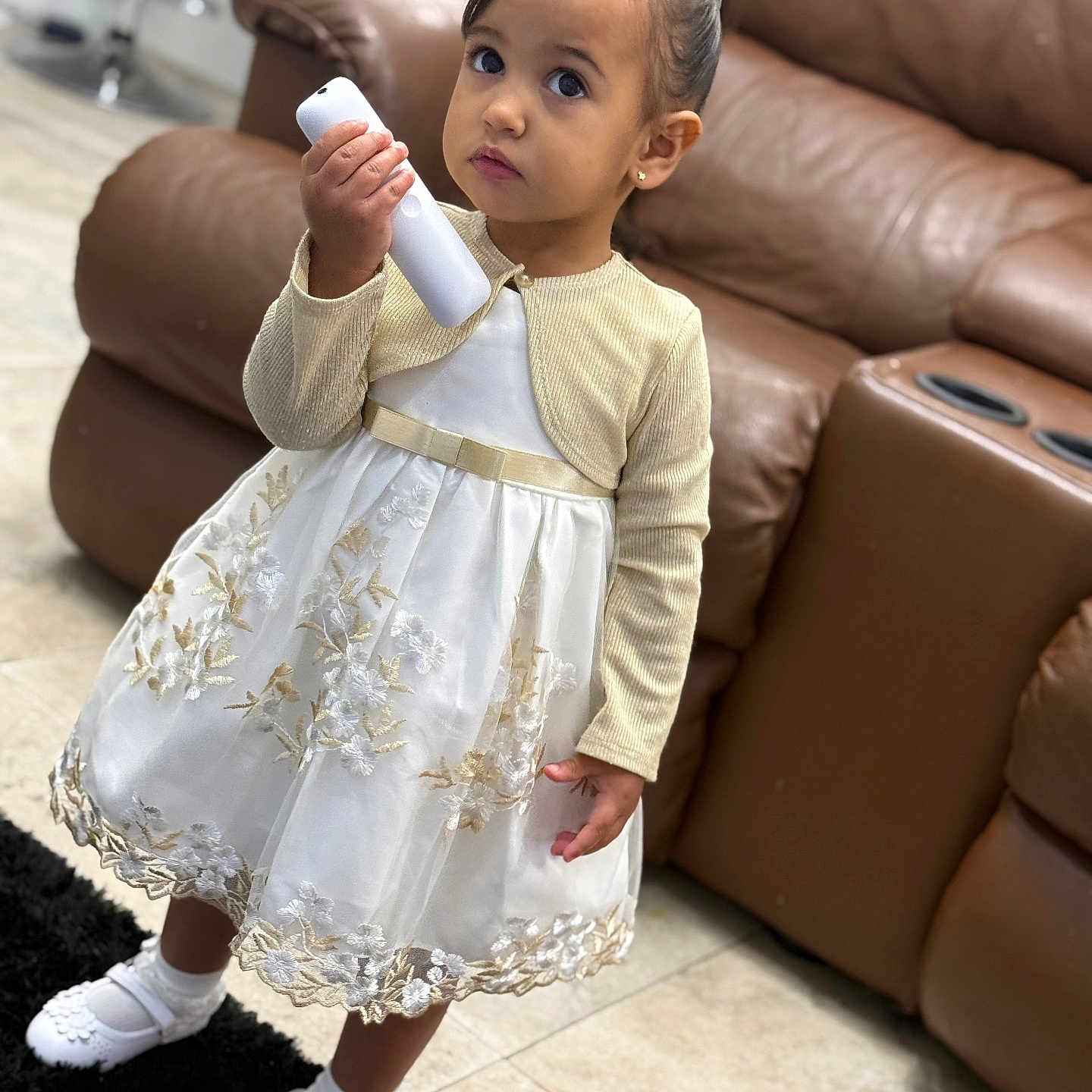 Brithanie is registered to the contest to win money with this photo: brown_sofa, carpet, child, curious, cute, dress, floor, girl, gold, indoor, living_room, person, portrait, remote_control, shoes, small, socks, standing, white, young