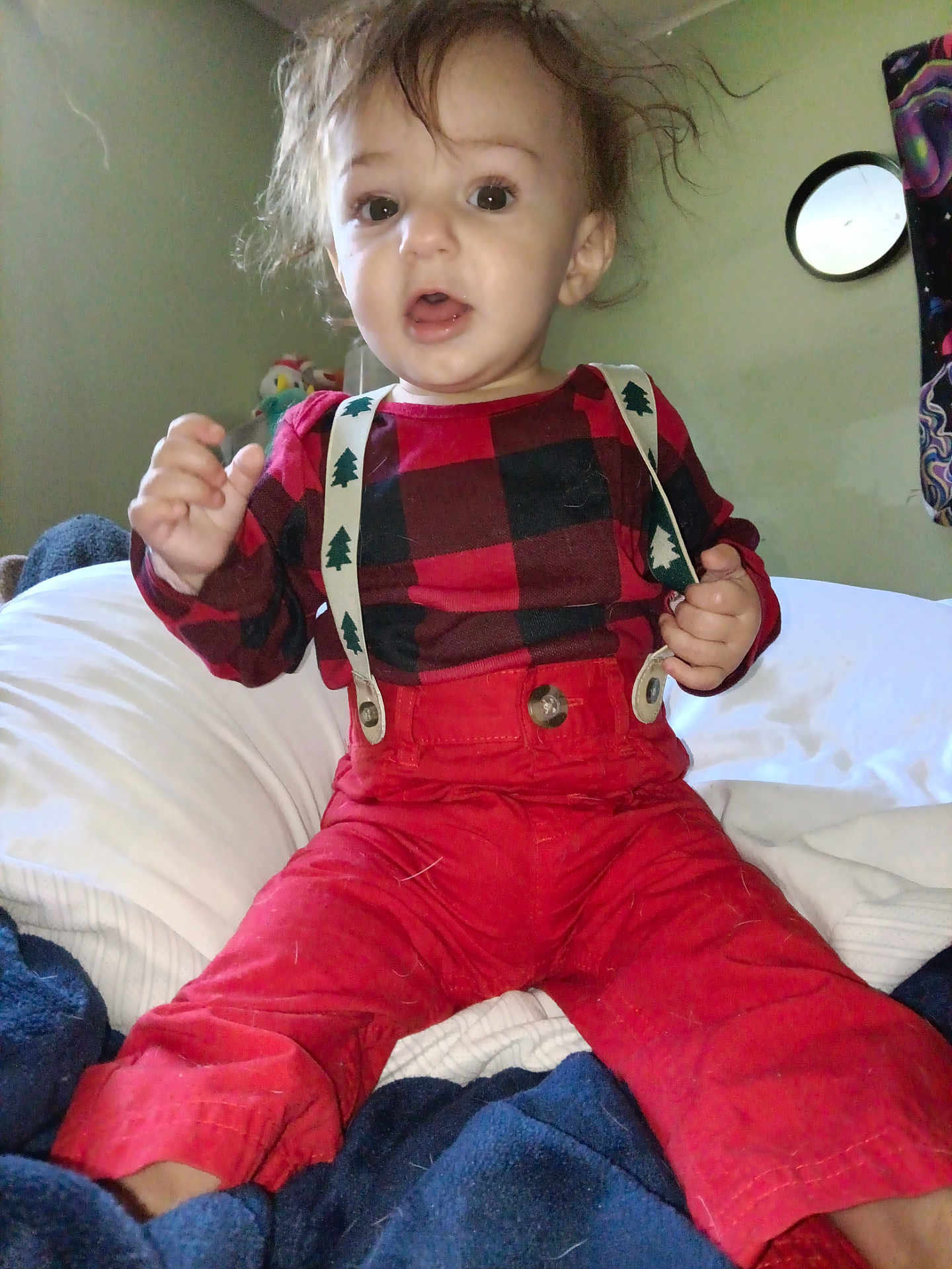 Atticus is registered to the contest to win money with this photo: toddler, child, red_pants, suspenders, checkered_shirt, bed, blanket, indoor, curly_hair, expression, face, person, clothing, sitting, cozy, home, furniture, young_child, cute, portrait