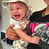 baby, child, smiling, laughing, hat, dinosaur_print, shorts, tshirt, adult_hand, beaded_necklace, indoor, laundry_basket, clothes, happy, person, holding, cute, face, portrait, home
