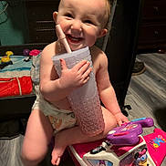 Ruby is registered to the contest to win money with this photo: baby, diaper, toy, toy_plane, cup, straw, smile, happy, indoor, floor, child, play, plastic, furniture, person, pink, purple, wood_floor, face, cute