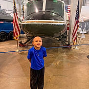 Xander joined the competition — help win amazing prizes! child, boy, helicopter, museum, american_flag, blue_shirt, black_pants, sneakers, indoor, vehicle, standing, smile, floor, rope_barrier, display, person, young, head, pose, casual_clothing
