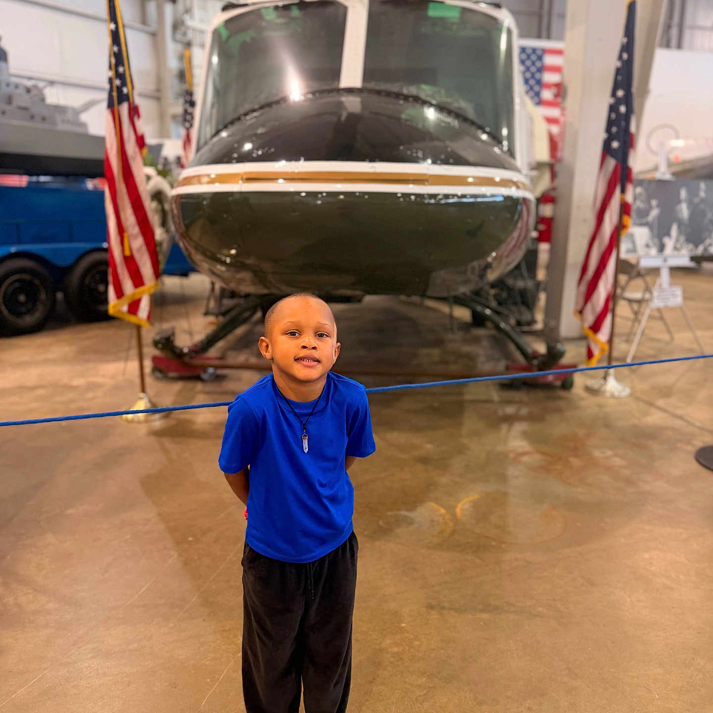 Xander joined the competition — help win amazing prizes! child, boy, helicopter, museum, american_flag, blue_shirt, black_pants, sneakers, indoor, vehicle, standing, smile, floor, rope_barrier, display, person, young, head, pose, casual_clothing