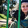 dog, black_dog, person, woman, green_dress, fence, outdoor, nature, sunlight, portrait, grass, trees, daylight, pet, animal, close_up, friendship, relaxing, calm, park