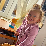 Léa participe au concours pour gagner de l'argent avec cette photo : toddler, child, smiling, curly_hair, pink_shirt, animal_print, floral_pants, wooden_chair, indoor, window, curtains, toy, table, natural_light, happy, person, face, blue_eyes, hair_bun, cozy
