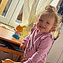 toddler, child, smiling, curly_hair, pink_shirt, animal_print, floral_pants, wooden_chair, indoor, window, curtains, toy, table, natural_light, happy, person, face, blue_eyes, hair_bun, cozy