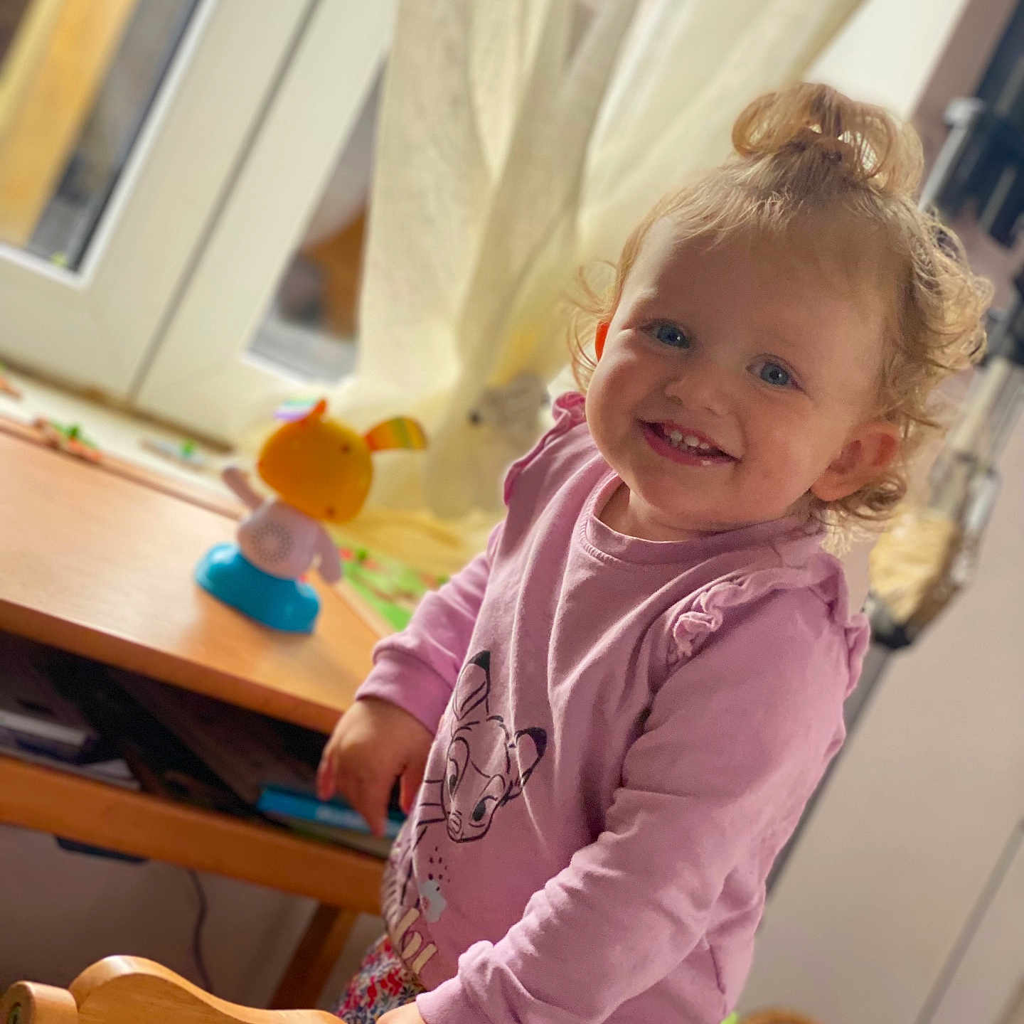 Léa participe au concours pour gagner de l'argent avec cette photo : animal_print, blue_eyes, child, cozy, curly_hair, curtains, face, floral_pants, hair_bun, happy, indoor, natural_light, person, pink_shirt, smiling, table, toddler, toy, window, wooden_chair