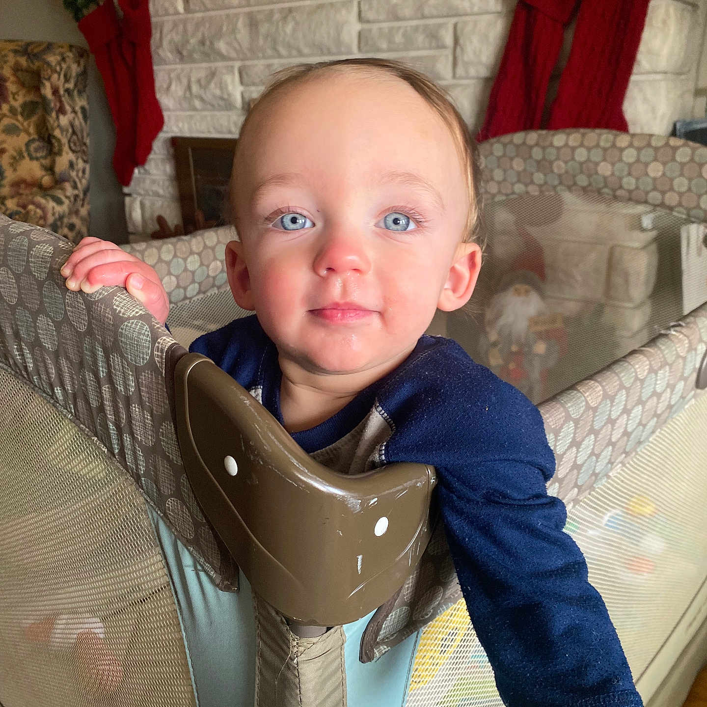 Nicholas is registered to the contest to win money with this photo: toddler, child, playpen, blue_eyes, rosy_cheeks, fireplace, stockings, holiday_decor, indoors, curious, face, hand, clothing, baby, home, furniture, smile, person, portrait, cozy
