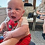 baby, child, person, indoor, waiting_room, chair, adult, overalls, red_clothing, hand, carpet, bag, people, seated, background, door, sign, blur, holding, casual_clothing