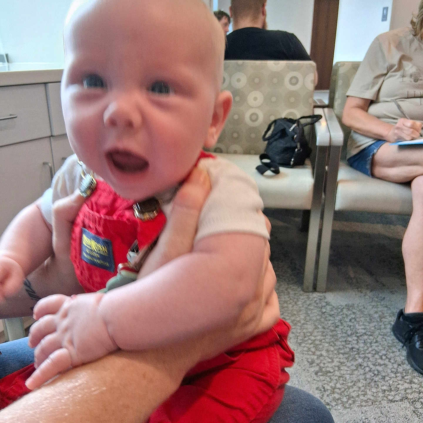 Grayson is registered to the contest to win money with this photo: adult, baby, background, bag, blur, carpet, casual_clothing, chair, child, door, hand, holding, indoor, overalls, people, person, red_clothing, seated, sign, waiting_room