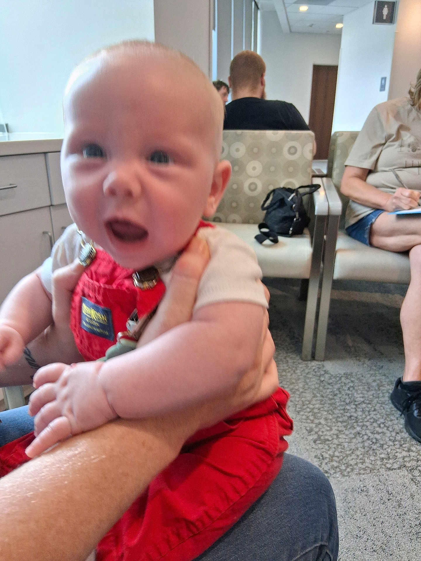 Grayson is registered to the contest to win money with this photo: baby, child, person, indoor, waiting_room, chair, adult, overalls, red_clothing, hand, carpet, bag, people, seated, background, door, sign, blur, holding, casual_clothing