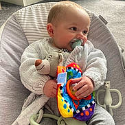 Justin is registered to the contest to win money with this photo: baby, pacifier, knitted_sweater, plush_toy, colorful_toy, baby_chair, carpet, pet_bed, indoor, child, cute, infant, cozy, smiling, hand, face, person, comfort, soft_toys, casual