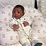 baby, infant, white_clothing, pillow, teddy_bear_pattern, bed, blanket, footwear, tiny_shoes, head, face, dark_skin, curly_hair, cute, indoors, soft_lighting, portrait, newborn, lying_down, resting