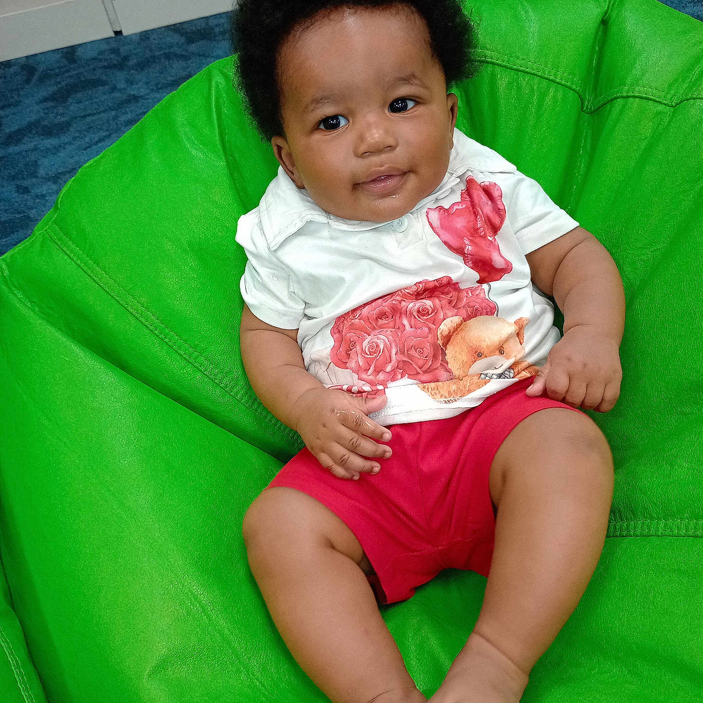 Josiah is registered to the contest to win money with this photo: baby, bean_bag, carpet, child, curly_hair, cute, furniture, green, happy, home, indoor, person, portrait, red_shorts, relaxed, roses, sitting, smiling, teddy_bear, white_shirt