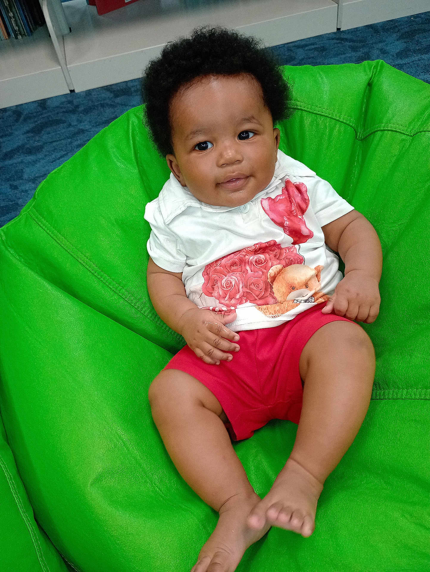 Josiah is registered to the contest to win money with this photo: baby, child, indoor, bean_bag, green, curly_hair, smiling, white_shirt, red_shorts, teddy_bear, roses, furniture, carpet, sitting, happy, cute, person, portrait, relaxed, home