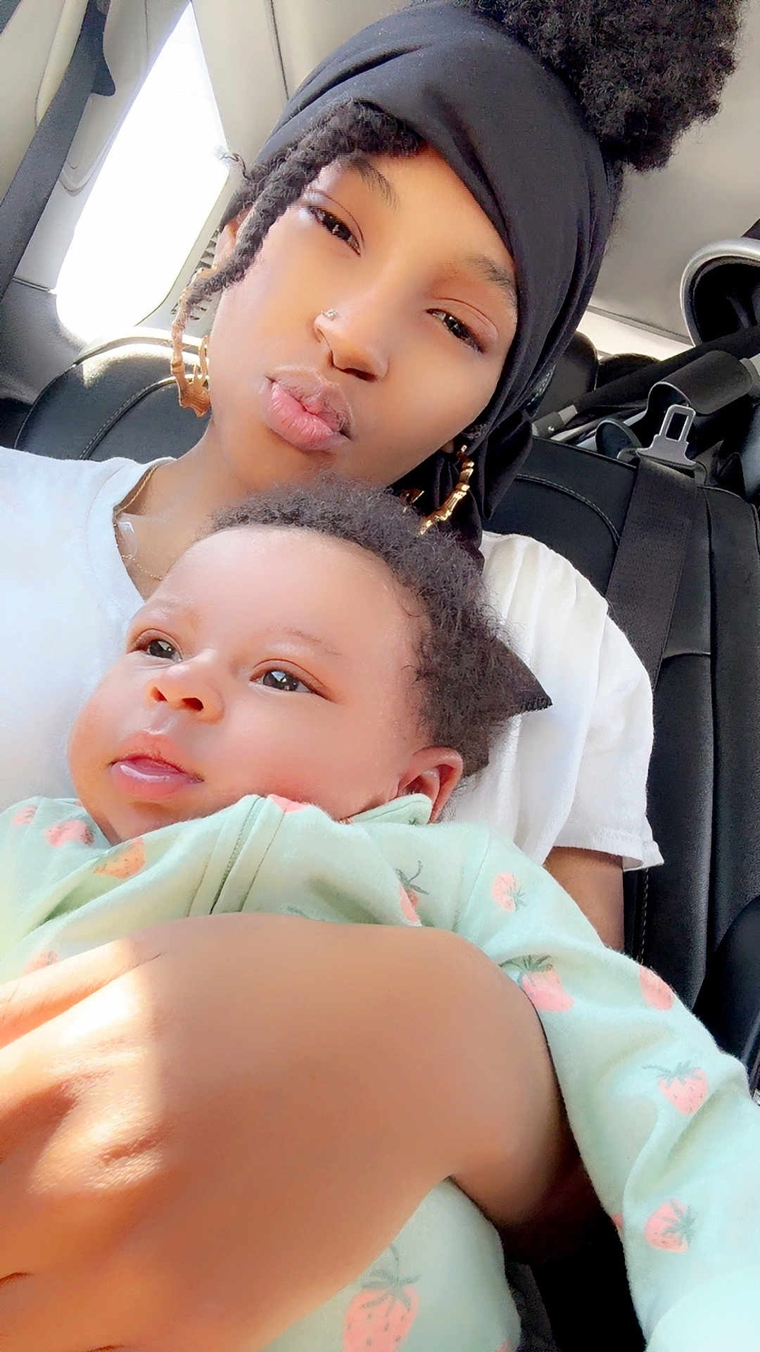 Nyari is registered to the contest to win money with this photo: woman, baby, child, car_interior, headwrap, earrings, onesie, strawberry_pattern, natural_light, holding, portrait, face, skin, hand, seatbelt, seat, window, black_hair, expression, closeup