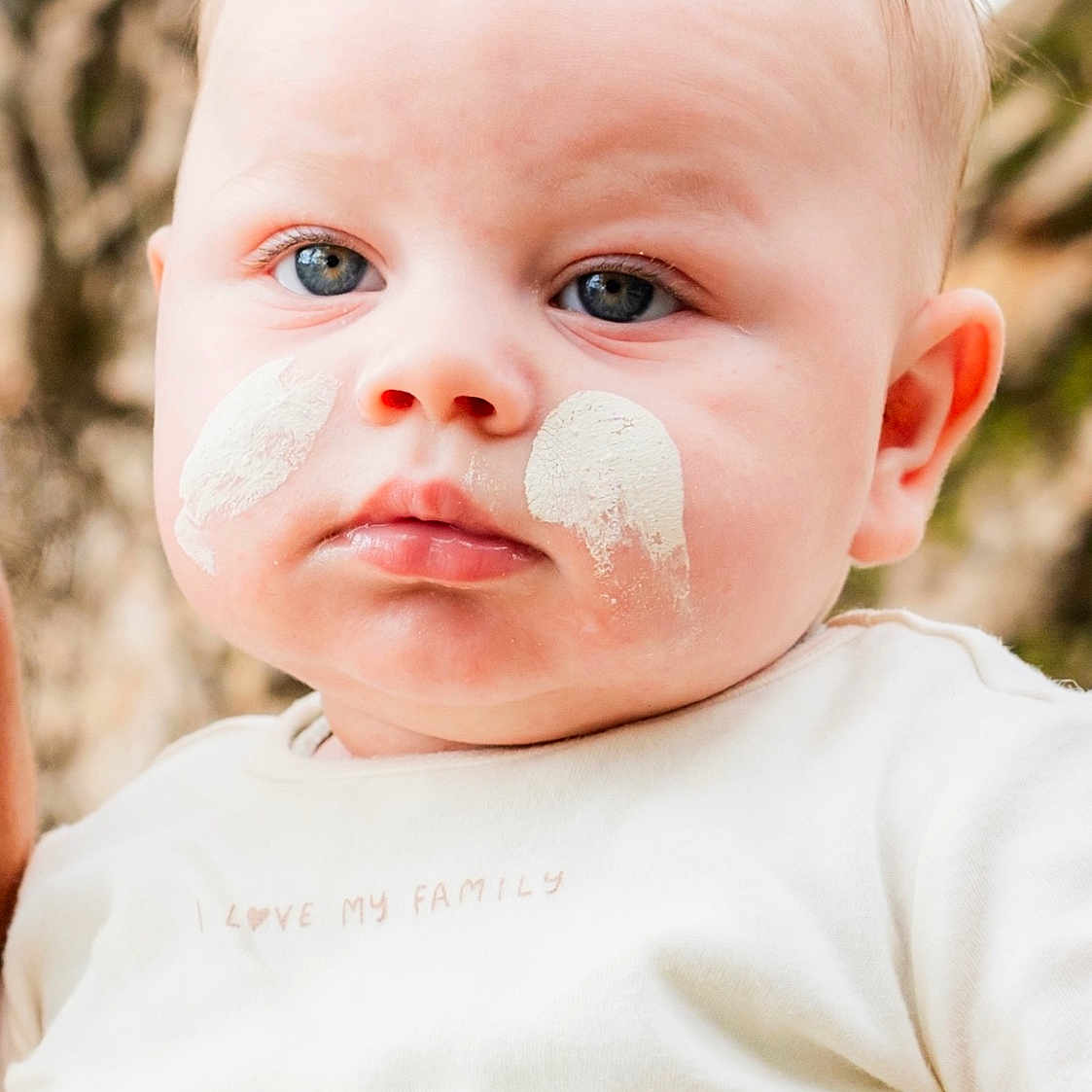 Jarrah is registered to the contest to win money with this photo: baby, blue_eyes, cheeks, child, close_up, cream, cute, expressionless, face, human, infant, natural_light, outdoor, person, portrait, skin, soft_focus, text_on_clothing, white_shirt, young_child