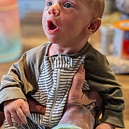 Zimri joined the competition — help win amazing prizes! baby, infant, child, person, hand, tattoo, clothing, striped_onesie, expression, wide_eyes, indoor, blurred_background, table, container, skin, head, face, holding, sleeve, zipper