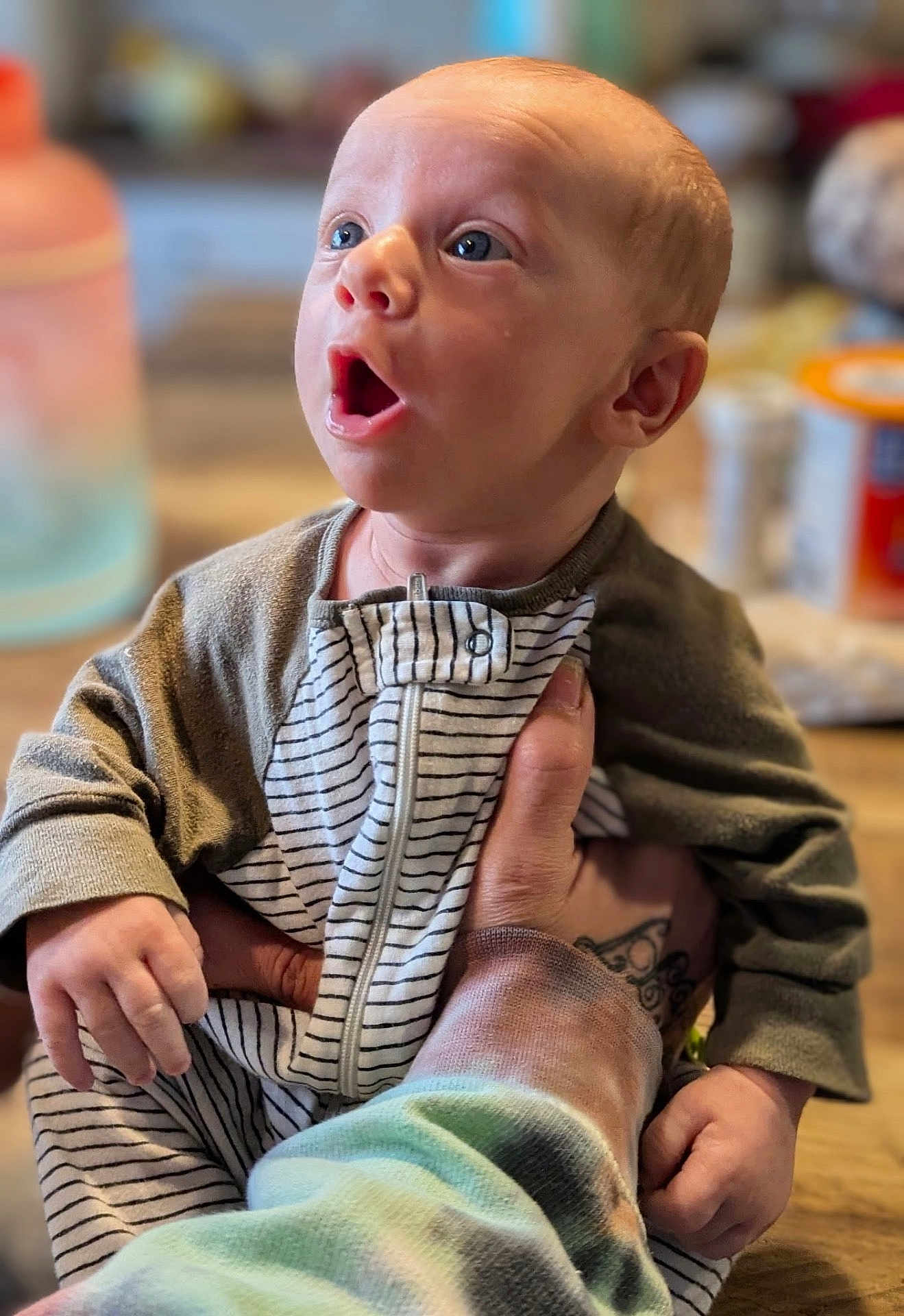 Zimri joined the competition — help win amazing prizes! baby, infant, child, person, hand, tattoo, clothing, striped_onesie, expression, wide_eyes, indoor, blurred_background, table, container, skin, head, face, holding, sleeve, zipper