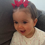 River is registered to the contest to win money with this photo: baby, child, face, smiling, happy, hair, pigtails, red_bow, onesie, heart_pattern, cushion, seated, indoor, cute, young_child, portrait, expression, infant, cheerful, clothing