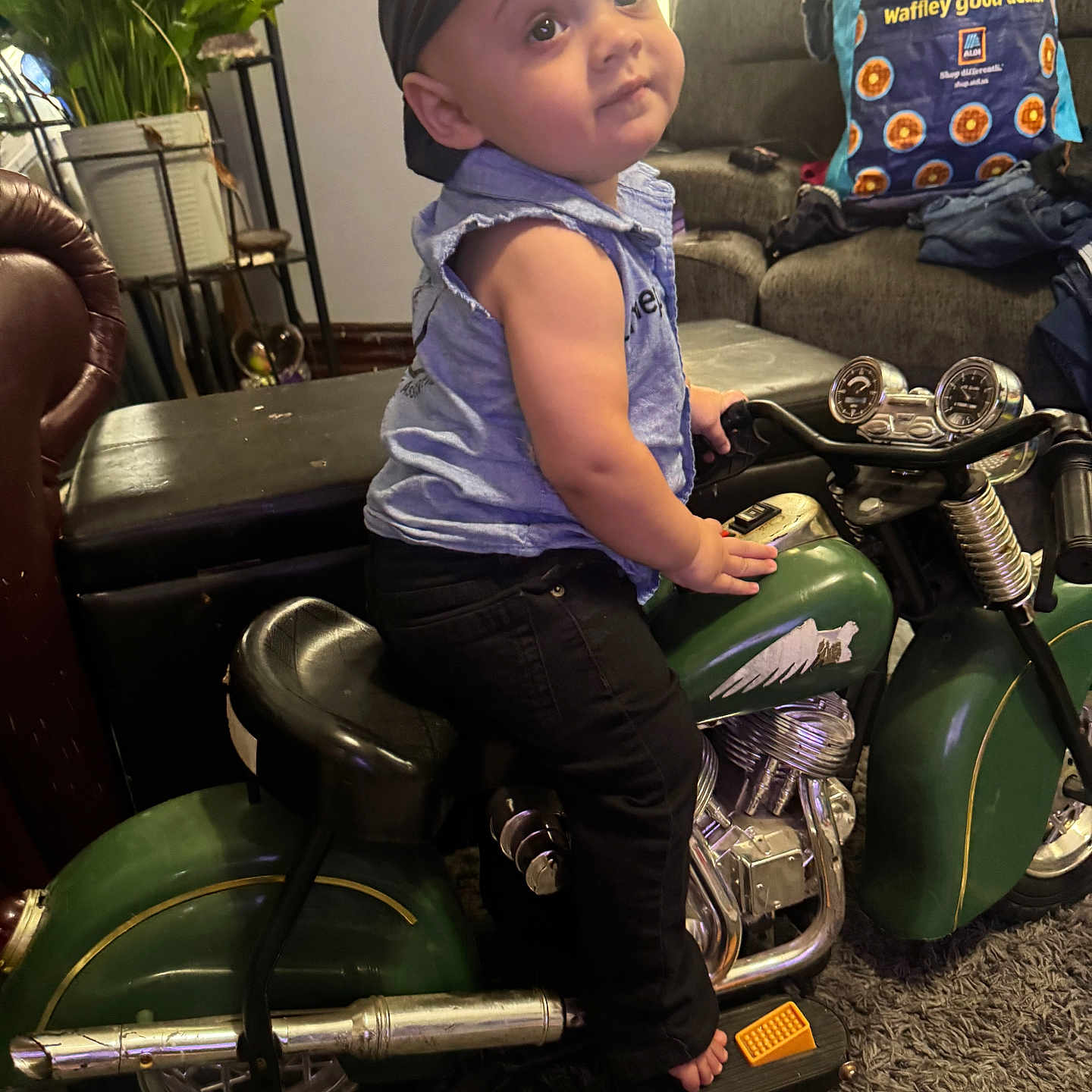 Zion is registered to the contest to win money with this photo: toddler, child, indoor, toy_motorcycle, headscarf, curious_expression, living_room, sofa, plant, carpet, furniture, clothing, barefoot, footrest, metal, green, black_pants, blue_shirt, face, person