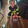 child, toy, firefighter_costume, fire_truck, wooden_floor, indoor, blurry, couch, furniture, person, playing, young_child, costume, pretend_play, home, living_room, curly_hair, barefoot, floor, kid