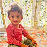 Kushin is registered to the contest to win money with this photo: toddler, child, sitting, bed, striped_shirt, green_pants, barefoot, floral_curtains, floral_bedspread, indoors, cute, smiling, portrait, baby, happy, person, cozy, home, young_child, casual_clothing