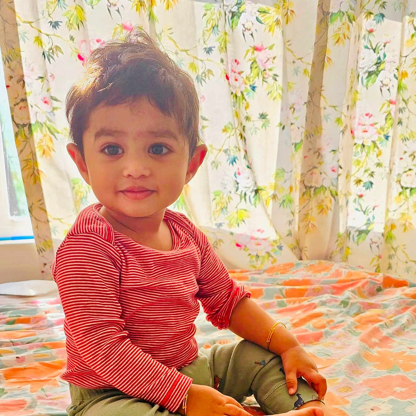 Kushin is registered to the contest to win money with this photo: toddler, child, sitting, bed, striped_shirt, green_pants, barefoot, floral_curtains, floral_bedspread, indoors, cute, smiling, portrait, baby, happy, person, cozy, home, young_child, casual_clothing