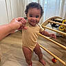 baby, child, smiling, curly_hair, onesie, wooden_floor, hand_holding, play_structure, toy, indoors, curtains, cheerful, standing, person, floor, light, room, support, cute, happy
