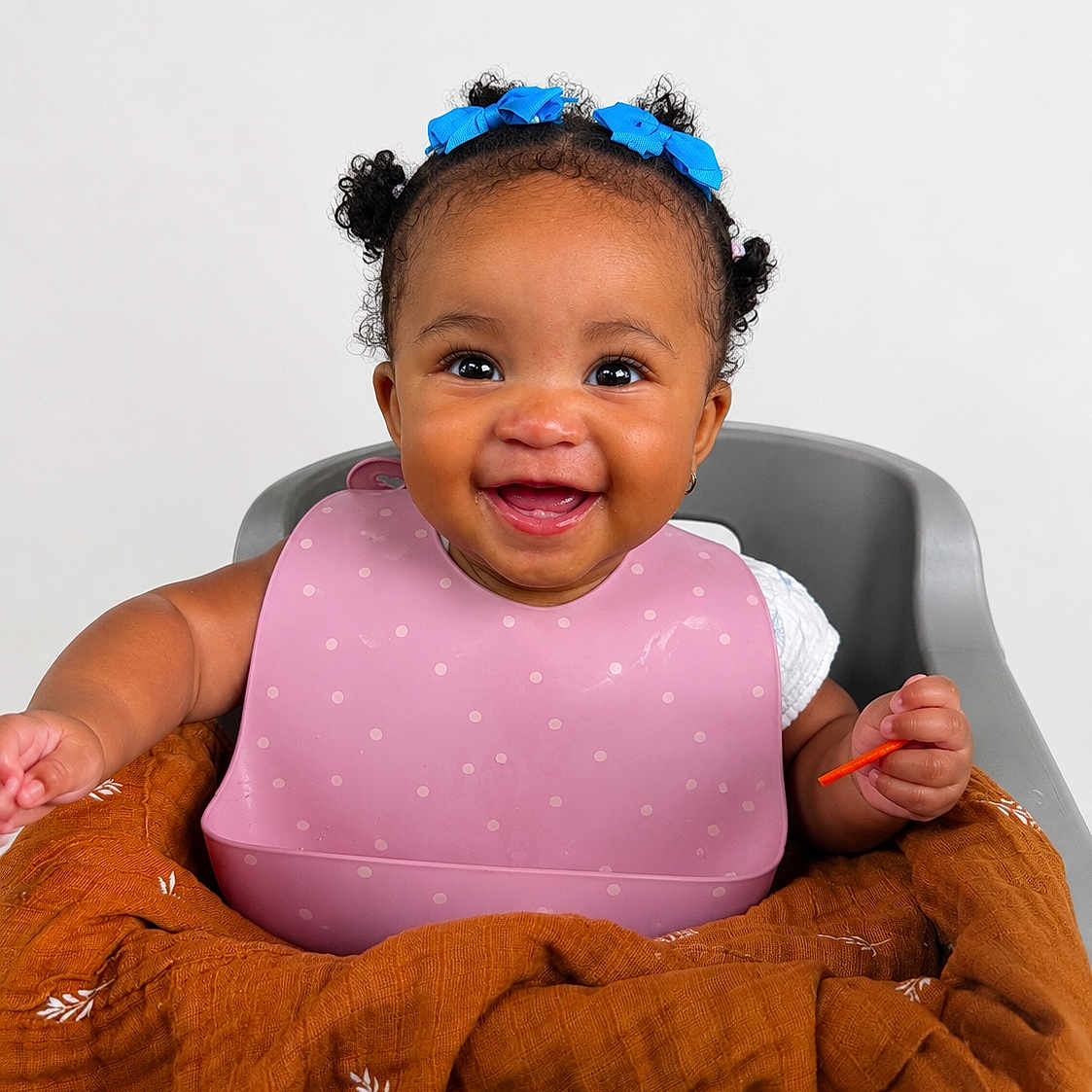 Ocean is registered to the contest to win money with this photo: baby, child, high_chair, bib, smile, happy, curly_hair, blue_bows, blanket, orange_blanket, teeth, cute, infant, person, indoor, portrait, clothing, sitting, face, young_child