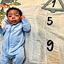 Ezra is registered to the contest to win money with this photo: baby, infant, child, blue_onesie, nike_logo, blanket, patterned_blanket, numbers, curly_hair, lying_down, indoor, cute, portrait, small_hand, small_foot, face, eyes, clothing, soft_fabric, young