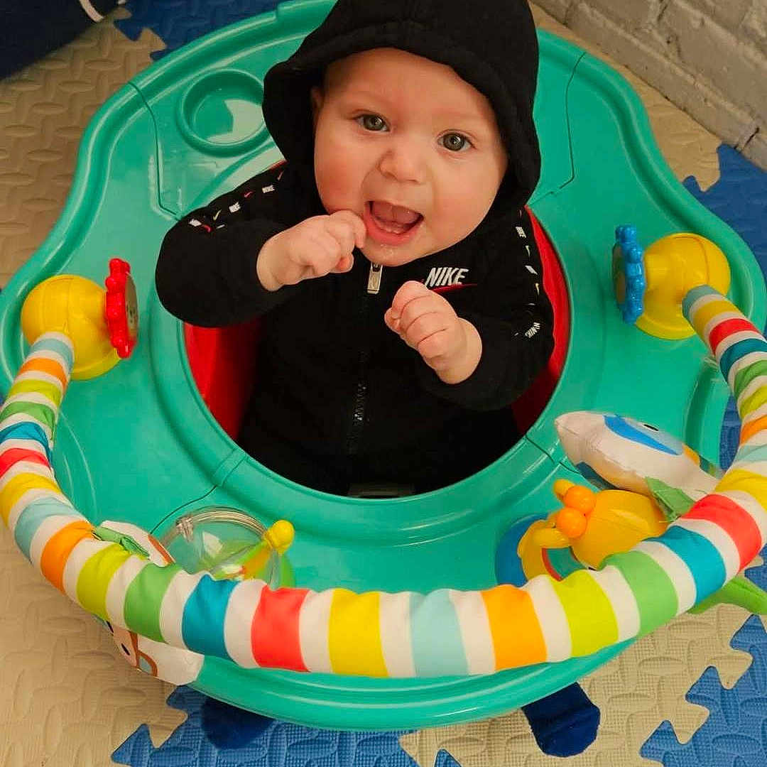 Kato joined the competition — help win amazing prizes! baby, child, hoodie, black_clothing, activity_center, toy, play_mat, colorful, happy, smiling, indoor, floor, plastic, person, infant, cute, seated, playful, bright_colors, young_child