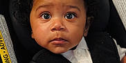Reginald is registered to the contest to win money with this photo: toddler, child, baby, car_seat, curly_hair, white_shirt, harness, seatbelt, safety, cute, portrait, indoor, young_child, person, face, hand, expression, black_harness, closeup, secure