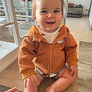 Tyam participe au concours pour gagner de l'argent avec cette photo : baby, child, smile, hoodie, orange_clothing, sitting, table, hand, indoor, wooden_table, happy, face, person, short_hair, skin, floor, kitchen, cabinet, casual_clothing, tooth