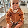 baby, child, smile, hoodie, orange_clothing, sitting, table, hand, indoor, wooden_table, happy, face, person, short_hair, skin, floor, kitchen, cabinet, casual_clothing, tooth