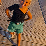 Brianna a rejoint le concours — aidez-le/la à gagner de superbes lots ! belt, black_shirt, blonde_hair, bracelet, casual_clothing, child, confident, crowd, evening, footwear, girl, happy, legs, outdoor, people, shorts, smiling, standing, white_shoes, wooden_deck