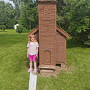 Gracey joined the competition — help win amazing prizes! barbie_tshirt, casual_clothing, child, church, daylight, girl, grass, greenery, miniature, nature, outdoor, pine_cone, pink_shorts, playful, smiling, standing, summer, tree, wooden_structure, young