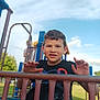 boy, casual_clothing, child, clouds, daylight, fence, fun, grass, happy, leisure, metal, nature, outdoor, park, person, play, playground, recreation, sky, smile