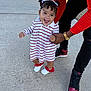 adult_hand, bows, casual_wear, child, concrete, curly_hair, dress, happy, outdoor, person, pigtails, red_accents, shoes, sidewalk, smiling, striped_clothing, supporting, toddler, walking, white_shoes
