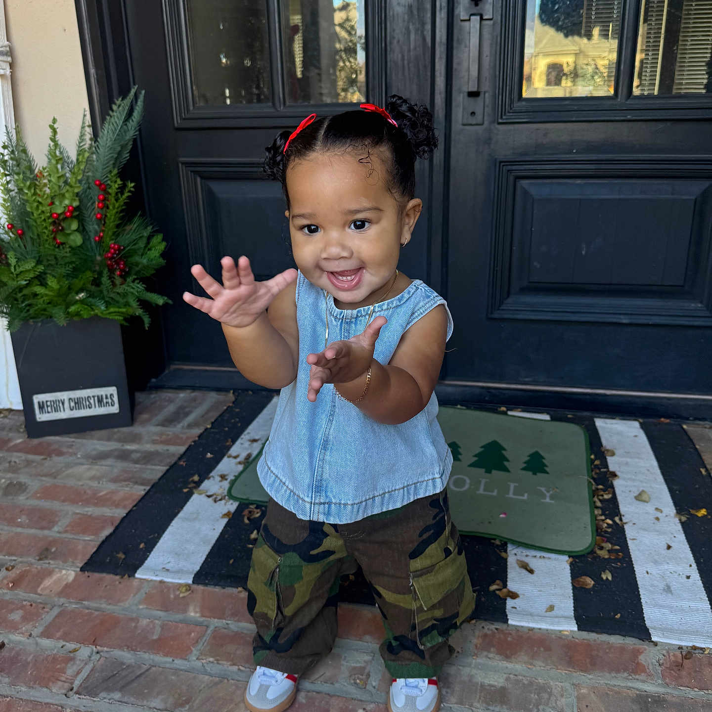 Khi is registered to the contest to win money with this photo: brick_porch, camouflage_pants, child, christmas_decor, curly_hair, cute, denim_top, front_door, happy, holiday_sign, outdoor, person, plants, portrait, reaching_hands, red_bows, smiling, sneakers, toddler, young_child
