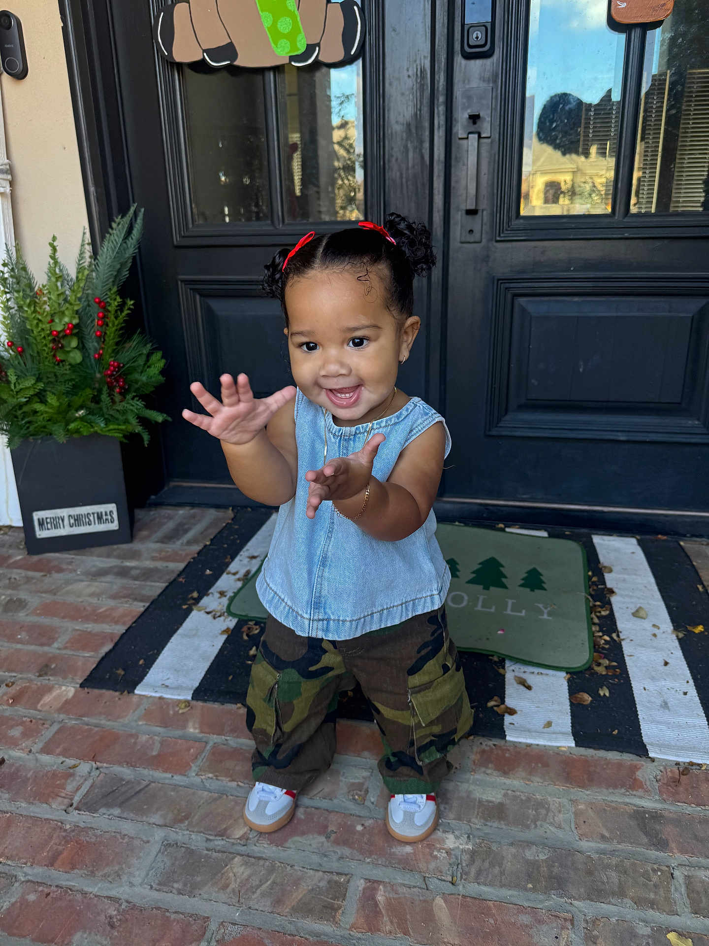 Khi is registered to the contest to win money with this photo: brick_porch, camouflage_pants, child, christmas_decor, curly_hair, cute, denim_top, front_door, happy, holiday_sign, outdoor, person, plants, portrait, reaching_hands, red_bows, smiling, sneakers, toddler, young_child