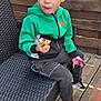 Ethan participe au concours pour gagner de l'argent avec cette photo : child, boy, green_jacket, black_pants, sneakers, outdoor_chair, wooden_fence, wooden_deck, snack, chocolate, pacifier, sitting, casual_clothing, daylight, expression, face, hand, footwear, leisure, outdoor