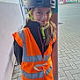 child, helmet, orange_vest, reflective_clothing, outdoor, paved_ground, pole, smile, person, young, safety_gear, long_hair, building, urban, daylight, casual, portrait, standing, headwear, colorful_background