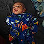 baby, infant, onesie, colorful_clothing, space_theme, blanket, lying_down, curious_expression, indoors, soft_lighting, face, hand, child, sleepwear, patterned_clothing, young_child, cozy, bed, resting, portrait