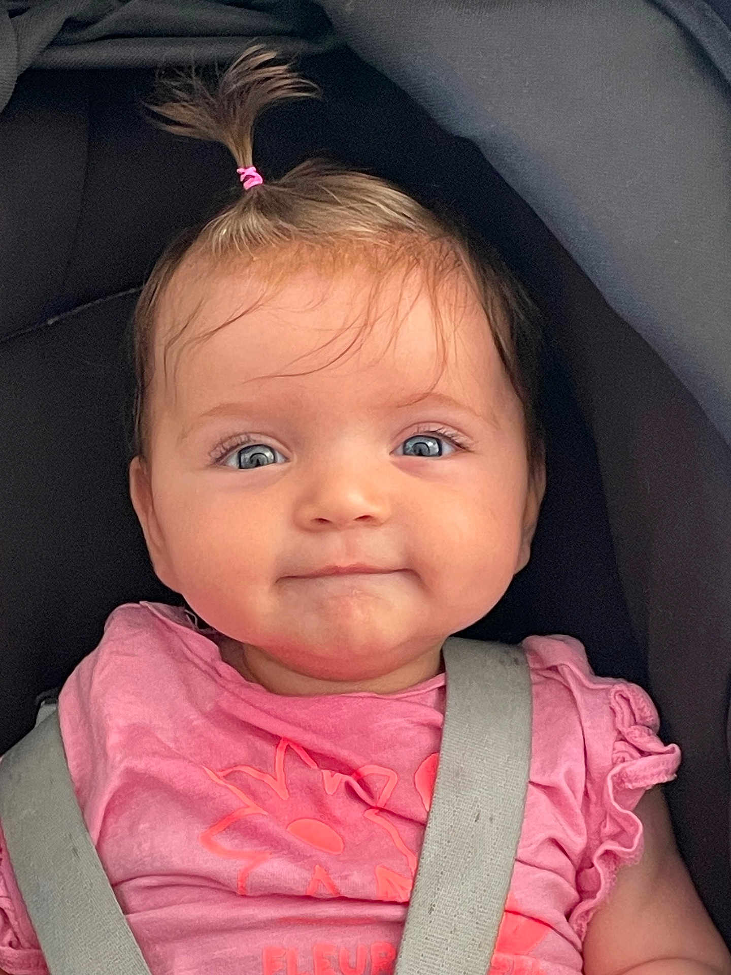 Ariane a rejoint le concours — aidez-le/la à gagner de superbes lots ! baby, child, girl, car_seat, pink_shirt, pony_tail, blue_eyes, smile, face, infant, clothing, seatbelt, cute, person, portrait, young_child, hair, expression, sitting, indoor